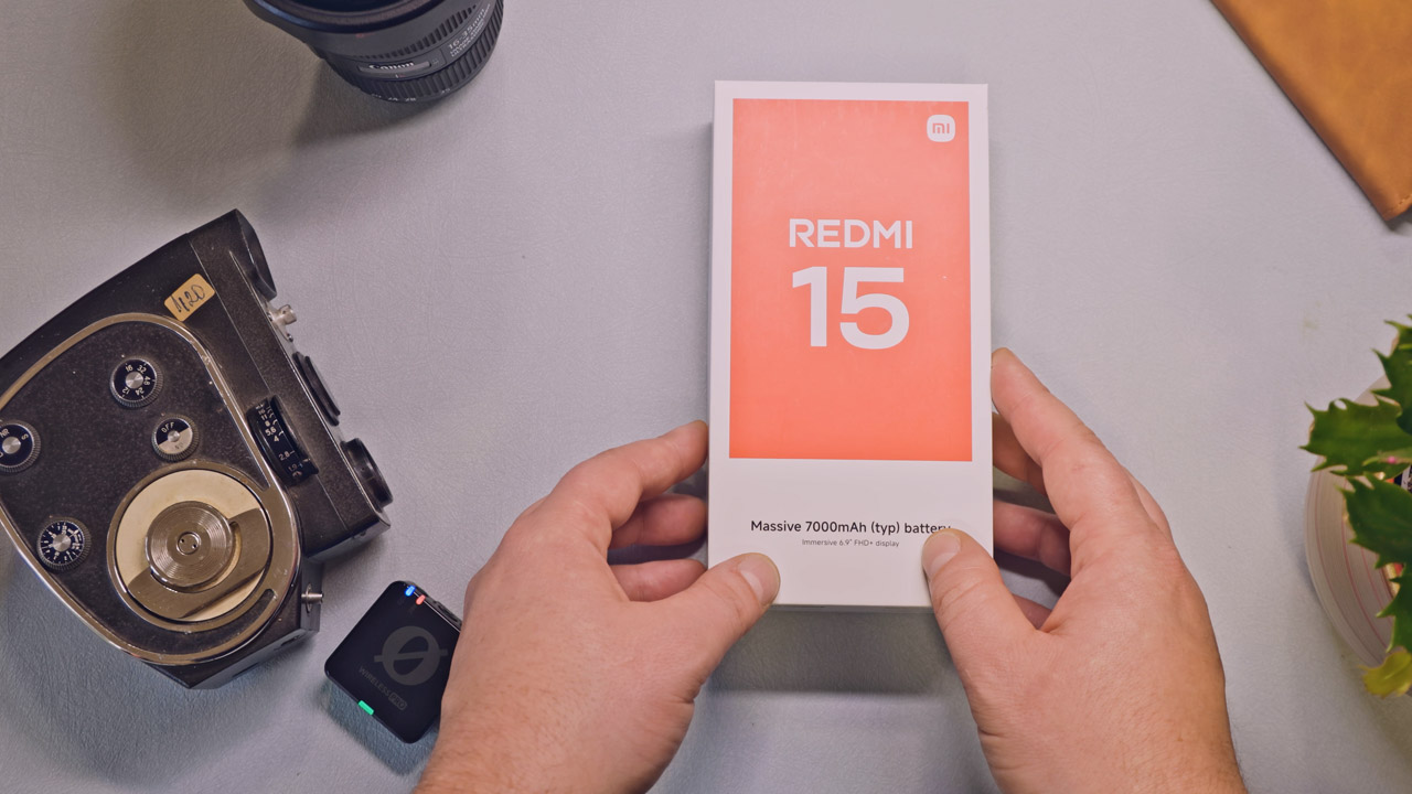 Gornja perspektiva koja prikazuje ruke i otvaranje kutije Xiaomi Redmi 15 uređaja na svijetlo plavoj pozadini.