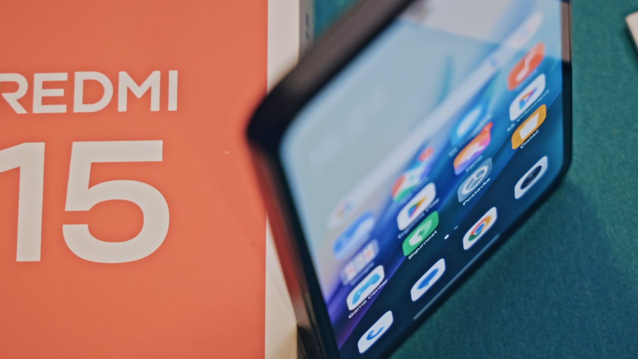 Krupni kadar s gornje perspektive Xiaomi 15 Redmi telefona na zelenoj pozadini gdje se vidi tekst i crveno bijela kutija dok telefon stoji uspravno.